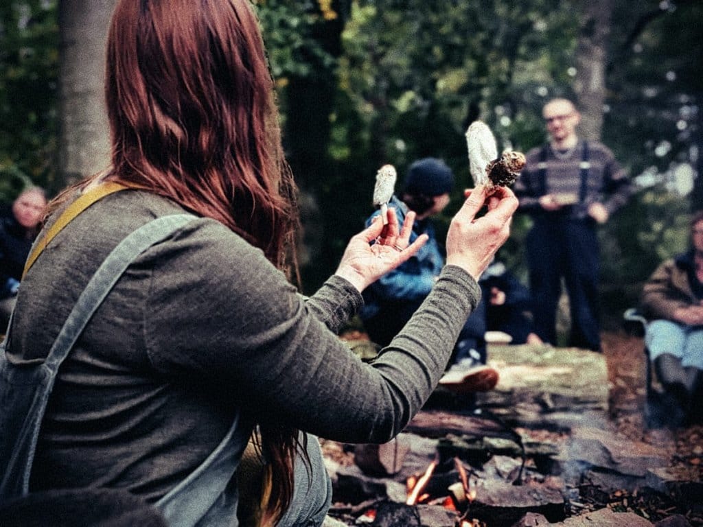 Bex holding up shaggy ink cap mushrooms by a campfire as a group looks on in the woods